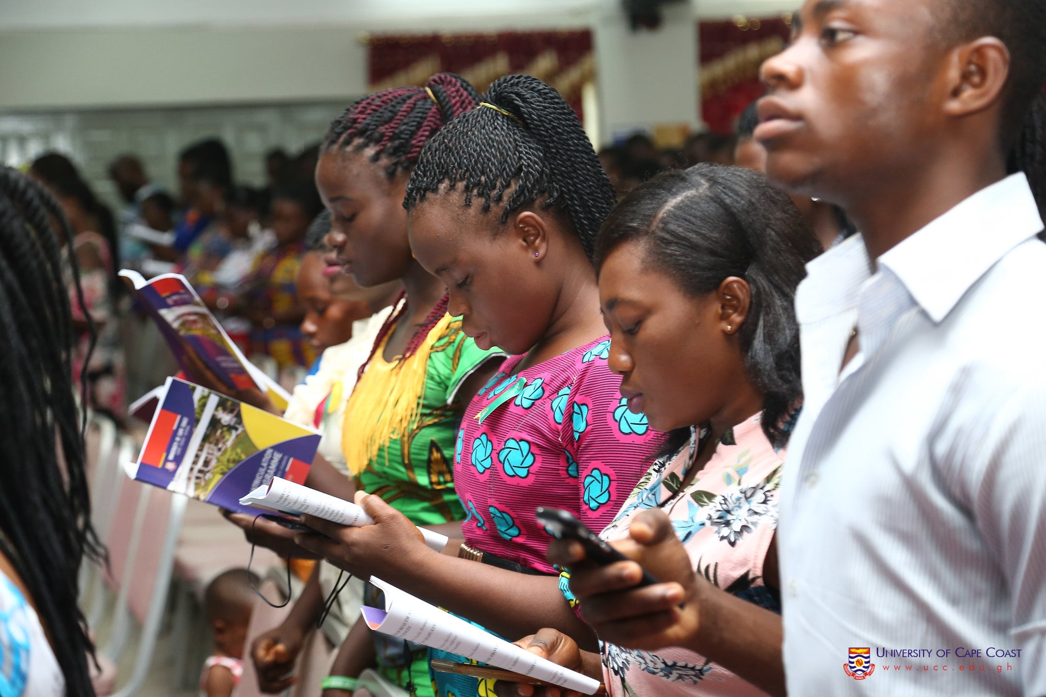 A group of students at matriculation holding books and notebooks. The students appear to be part of a diverse community, with various skin tones and hairstyles. Many of the students are wearing colorful traditional clothing patterns. Some are focused on reading materials while others are looking up attentively, suggesting they are engaged in learning or instructional activities. The setting appears to be an educational institution, as indicated by the University of Cape Coast logo visible in the bottom right corner of the image.