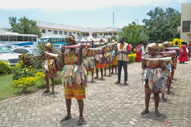 A group of students, likely performers or dancers, wearing vibrant traditional African attire. They are standing in a line, facing the camera, with their arms stretched out to the sides. Their clothing features colorful patterns and designs characteristic of various African cultures. The performers appear to be posing or preparing for a cultural performance or celebration. The setting is outdoors, with buildings and vehicles visible in the background, suggesting an urban or institutional location. The image captures a lively and festive cultural expression through dance and traditional dress.